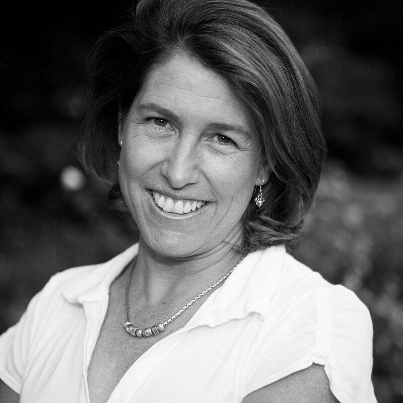 The image shows a black and white portrait of a smiling woman with short, wavy hair. She is wearing a white collared shirt and a necklace. The background is blurred, suggesting an outdoor setting with foliage. The woman appears to be in her late 30s or early 40s, and her expression is friendly and approachable.